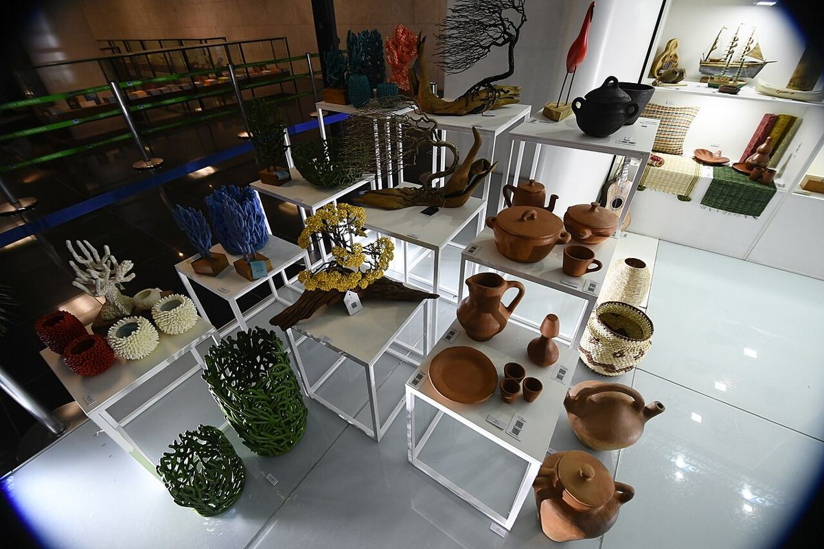 Panelas, jarras e pratos de cerâmica vermelha ao lado de cestos de palha e esculturas de madeira em uma exposição.