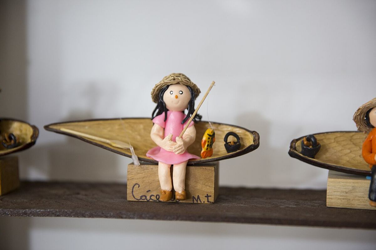 Boneca de chapéu de palha sentada em uma canoa feita de casca de semente, segurando uma vara de pescar com um peixe.
