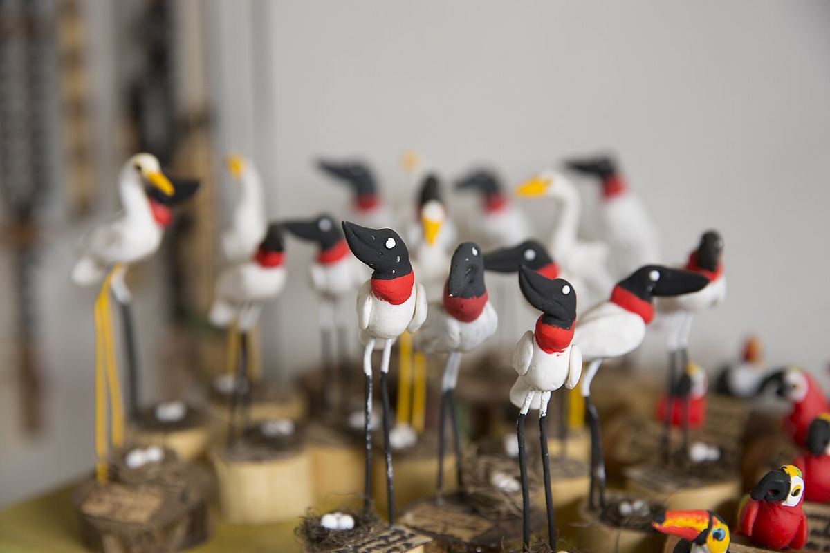 Dezenas de pequenos tuiuiús de barro pintados de branco, preto e vermelho, sobre bases de madeira.